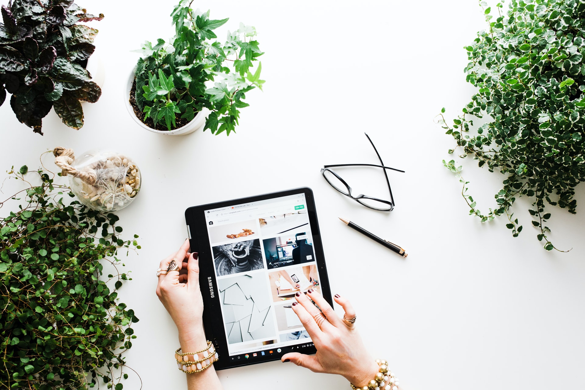 Hands using a tablet on a white desk, surrounded by plants, glasses, and a pen.